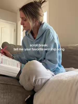 A woman with blonde hair, wearing a light blue hoodie and white sweatpants, sits on a grey couch, knees bent, holding a book with pink highlights. The image is well-lit, with soft, natural light, and a soft-focus background featuring a white door. Text overlayed on the image says, 'scroll break: tell me your favorite worship song'. Overall a calm and inviting scene.