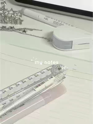 The image shows a close-up of study supplies arranged on a white surface. A white highlighter pen and a clear ruler with printed numbers and markings are the main elements. A transparent pen and a few metallic star-shaped decorative pieces partially visible on the upper left corner. The composition is minimalist with a focus on clean lines and a soft, neutral color palette, suggesting a study or work-related setting.