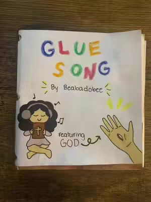 A hand-drawn illustration on a white paper, featuring the words "GLUE SONG" in colorful letters at the top. Below, the text "By Beabadoobee" and a drawing of a girl with headphones holding a book with a cross. The text "featuring GOD" points to a hand emerging from the right. The style is simple, childlike, and colorful.