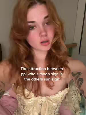 A woman is seen from the chest up, with long wavy auburn hair. She is looking directly at the camera. The image has text at the bottom of the frame. She's wearing a cream-colored corset.
