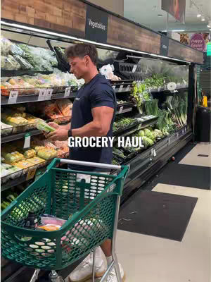 A man with short brown hair stands in front of refrigerator cases in a grocery store, holding a head of lettuce and looking at it. Text overlay in white font reads 'GROCERY HAUL'. A shopping cart filled with items is in the foreground.