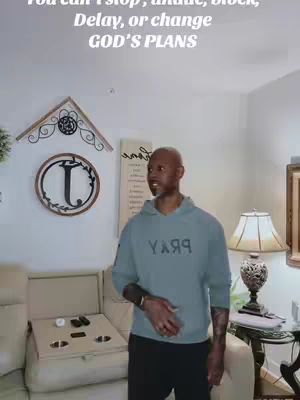 The image shows a man in a home setting. He is wearing a blue-green "Pray" hoodie and dark pants. The background has a beige couch, a white wall with framed decor, and a table with a lamp. Above the man, text overlays read, You can’t stop, undo, block, Delay, or change GOD’S PLANS.