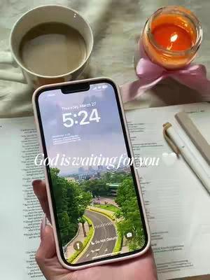 The image shows a phone displaying its lock screen, which has a scenic background with a road and greenery during the daytime. The phone is held by a person with a light skin tone. Next to the phone is an open book with text, a pen, and a candle, suggesting a study or devotional setting. The overall composition of the image is bright and uses a balanced arrangement of objects.
