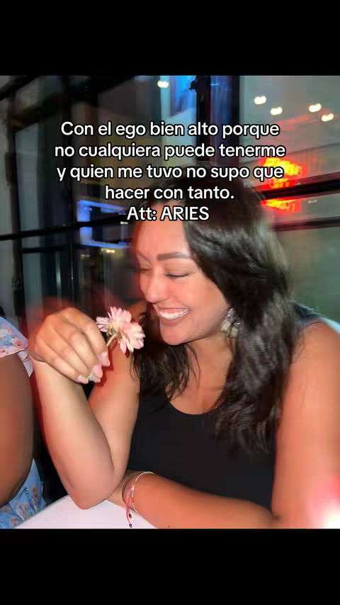 A selfie-style video cover image featuring a woman smiling subtly while holding a pink flower. She is indoors with a blurred background. Text in Spanish is overlaid on her image.