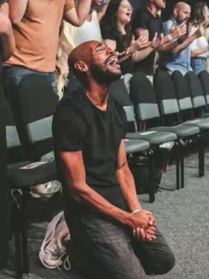 A man kneeling with his hands clasped in front of him, with a look of intense emotional expression. Behind him, a group of people stand and are raising their hands in worship.