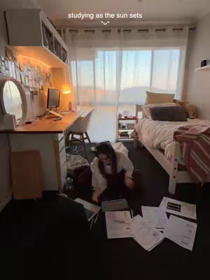 A woman is sitting on the floor surrounded by papers and a laptop working on a desk in a bedroom setting. "Studying as the sun sets" is written above the image. A bed is in the background.