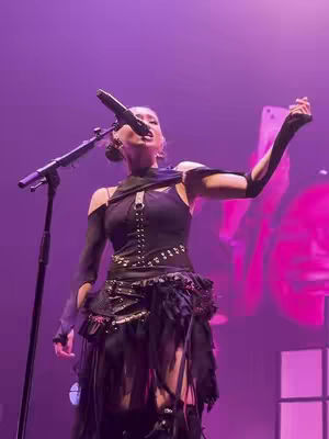 A woman with dark hair is singing into a microphone on stage, dressed in a black, edgy outfit with cut-outs. The background is a purple wash with some indistinct shapes, suggesting a concert setting.
