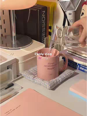 A cozy, well-lit study setup with a pink desk, a pink mug with 'I love strawberries much', books, a desk lamp, and various stationery items. The image captures a relaxed, inviting study environment.