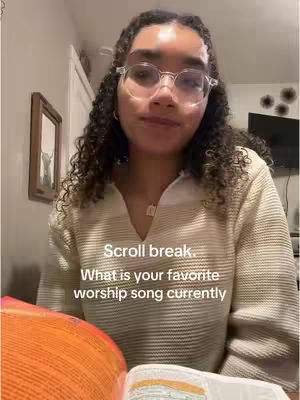 A young woman with curly hair and glasses is seated indoors. She is wearing a light colored sweater and the background includes a framed picture and a television. Text on the screen asks about favorite worship songs.