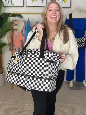 A woman with blonde hair, wearing a jean jacket and a pink shirt, holds a black and white checkered duffel bag. She's smiling with her mouth open. In the background are clothes on hangers, and a mannequin in a blue outfit.