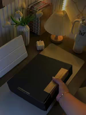 A well-lit, top-down shot of a desk space. The focus is on a black box sealed with tape, held by a hand with a bracelet. A Bible and a lamp are visible. The scene has a warm color palette.
