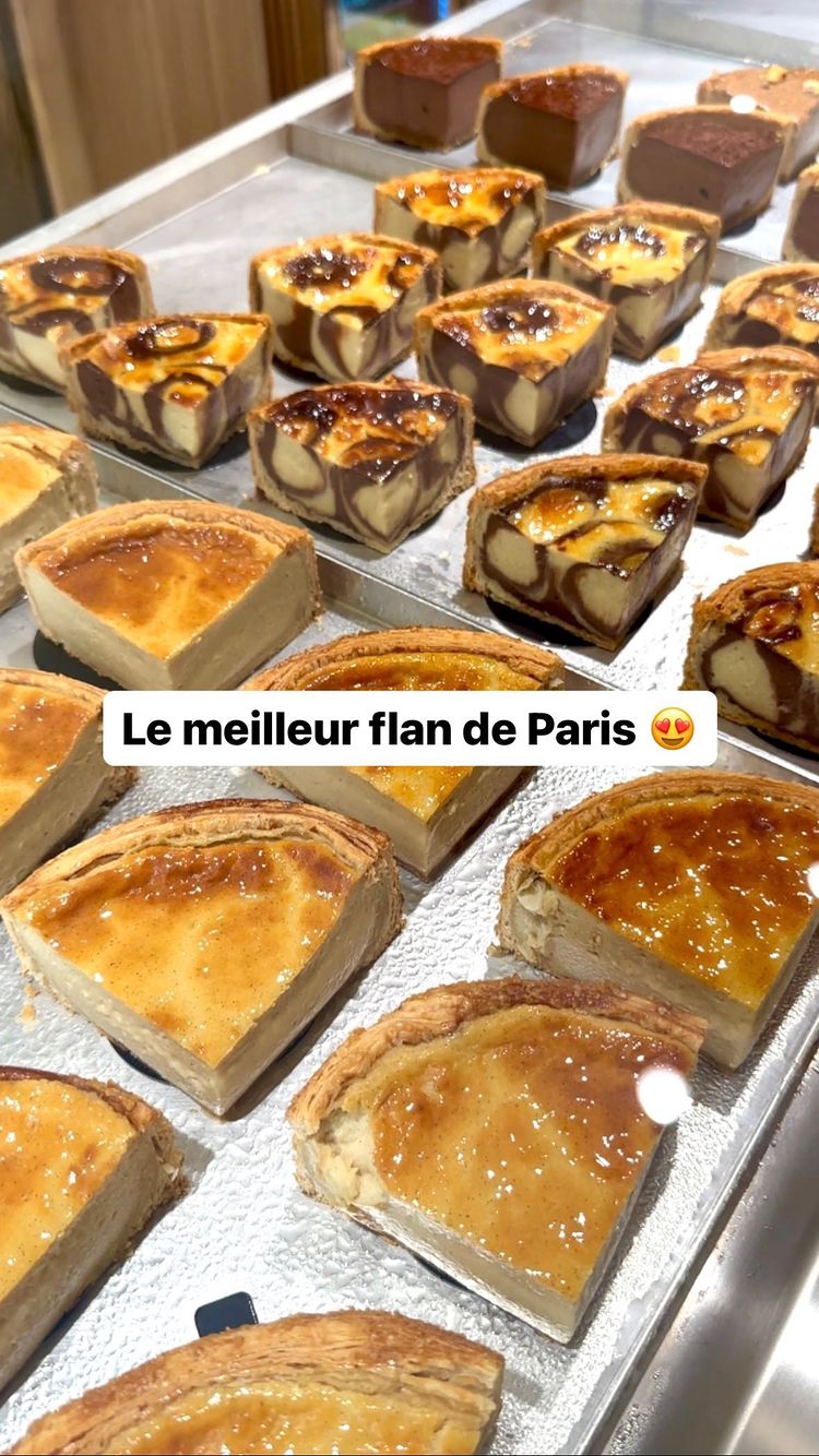 A photograph showcases a variety of flan pastries arranged on metal trays. The flans are presented in individual, square-shaped portions with a golden-brown, caramelized top crust. Some have a marbled appearance, indicating different flavor combinations, while others display a solid custard surface. The camera angle offers a direct, eye-level view, emphasizing the details of the pastries. The overall composition is well-lit, highlighting the texture and appeal of the flans, with the background setting appearing to be a shop display or kitchen.