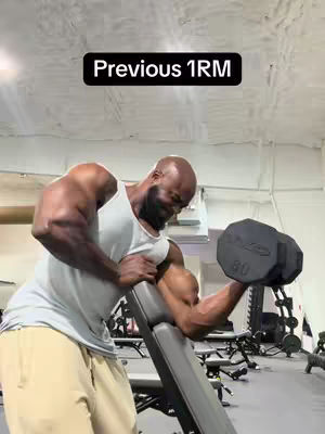 A muscular man, dressed in a white tank top and tan shorts, is shown from the waist up in a gym setting. He's mid-exercise, in the process of completing an arm curl while seated on a specialized bench. The background includes gym equipment and a concrete ceiling.