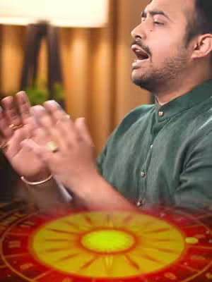 A man with a beard is clapping his hands in front of a stylized astrological wheel. He wears a green shirt and a gold ring. The background is blurred, showing warm lighting.