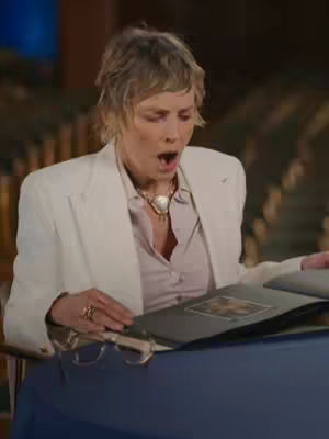 The cover image shows Sharon Stone with an open mouth, reacting to something she is looking at. She is wearing a white blazer, a light-colored blouse, and a necklace. She is seated at a table, and a book or photo album is open in front of her. The background is somewhat blurred but appears to be a room with decorative elements.