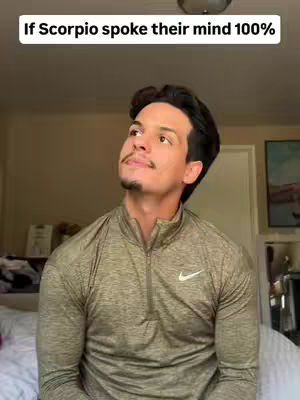 A man is looking upwards in a room. He is wearing a long-sleeved olive-green shirt. The man has dark hair and a small amount of facial hair. The background includes a light-colored wall, a bed, and a framed picture.