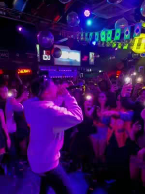 A dimly lit club scene with a man in a white hoodie dancing in the foreground. He is the focal point, with a crowd of people visible in the background, many holding up phones with lighted screens.
