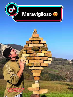 A man in casual clothing is balancing a complex tower of bricks outdoors with a scenic backdrop.
