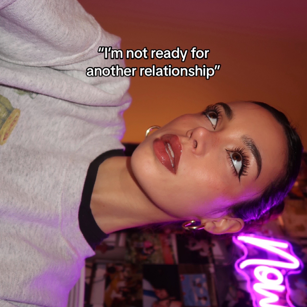 A young woman with dark hair and dramatic eye makeup is looking up, bathed in a blend of warm and cool lighting. A text overlay reads, "I'm not ready for another relationship." A neon sign is visible in the background.