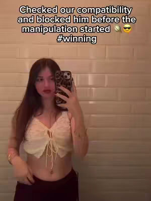 A young woman is taking a mirror selfie. The background shows a plain white tiled wall. The woman has long dark hair, and is wearing a cream top, and black pants. She holds a leopard print phone to take a selfie.