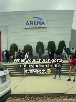 An exterior shot of an arena with numerous people gathered in front. The building is a light gray with a large sign that reads 'ARENA' and a smaller sign below naming a college park. Several green bushes and trees line the front steps. In front of the arena, individuals are standing and walking. A white car is visible in the bottom left corner.
