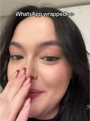 A close-up shot shows a woman with dark hair, fair skin, and a slight smile. She is holding her hand near her mouth, with her fingers lightly touching her lips. The nails are painted with a red/burgunndy polish. "WhatsApp wrapped >>" is written in black text.
