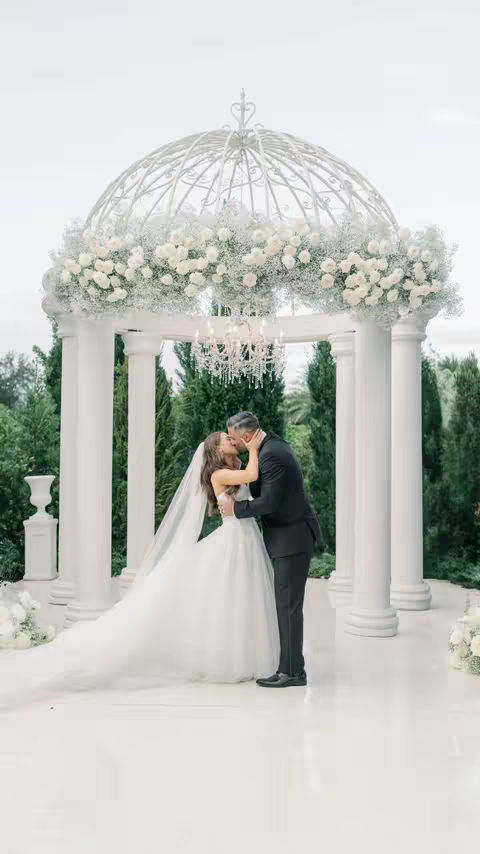 A photo of a wedding ceremony shows a couple kissing under a white gazebo adorned with white flowers. Lush green trees form the backdrop, with a white flooring, and a beautiful hanging chandelier.