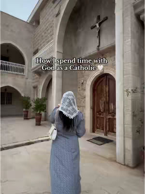 A woman in a long gray dress and white head covering stands facing away from the camera. She is in front of a large building with arched doorways and a cross. The text overlay reads, How I spend time with God as a Catholic with a white heart symbol.