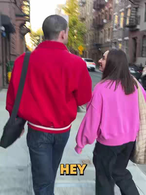 Two people are walking side-by-side on a city street, captured from a slightly low angle. The person on the left wears a red jacket and jeans. The person on the right wears a pink sweatshirt and black pants. The background shows buildings made of brown bricks.