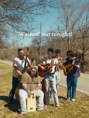 A group of five musicians in a casual outdoor setting, with a road and trees in the background. The band is composed of men with guitars, a bass, and various other instruments. The overall scene has a warm, natural aesthetic.