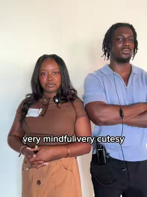 A medium shot of a man and woman standing side by side. The woman is on the left wearing a brown top and light tan skirt. She is wearing glasses around her neck and a lav mic. The man on the right is wearing a blue polo shirt and holding himself as if his arms are crossed. He also has a lav mic and dark pants.