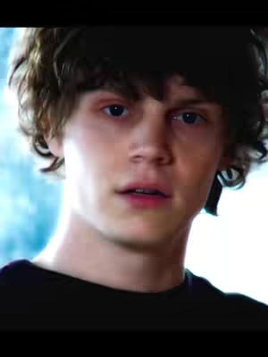 A close-up of a light-skinned man with short, wavy brown hair. He looks directly at the camera with a slight expression of surprise on his face. His lips are slightly parted, the lighting suggests a soft, natural setting, and the background indicates a blurred area outside the frame, possibly outdoors.