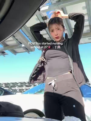 A woman with light skin and dark hair is seen outdoors, likely near a vehicle. She is wearing a gray top, black pants, and a black jacket she's lifting, possibly putting her hair up. Above her is a car's open sunroof or a similar structure. The background shows sky and sunlight, with faint glimpses of parked cars. Text overlay indicates a focus on her nursing school journey.