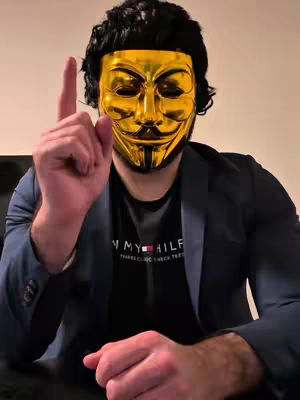 A person wearing a golden Guy Fawkes mask and a black wig raises their index finger, dressed in a black Tommy Hilfiger t-shirt and a dark blue blazer. The background is plain and the lighting is neutral.