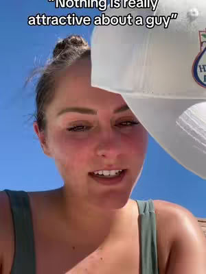 A woman with light skin and her hair pulled up in a bun is seen from a low angle against a bright blue sky. She is wearing a green top and a white hat partially blocks the view of the camera. The woman is looking at the camera and speaking. Text overlaid on the image reads ""Nothing is really attractive about a guy""