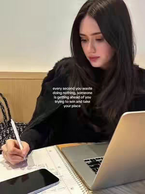 A woman with long black hair is seated, writing on a notepad with a pen. A laptop, a cell phone, and a notepad are on the table. The overlay text conveys a motivational message.