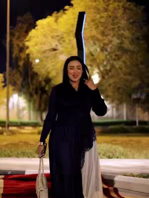 A woman in a dark dress and hijab stands outside at night, holding a silver purse. The background has blurred lights and trees; a decorative structure is prominent.