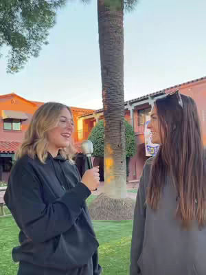 Two young women are standing outside, seemingly in a college setting. One is holding a microphone and speaking to the other, who stands listening. Buildings and trees are softly visible in the background.