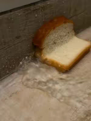 A partially submerged slice of white bread rests on a wet, wooden surface. The bread's texture is visible, with a golden-brown crust and a fluffy interior. Water surrounds the bread, creating a reflective surface and adding to the wet appearance. The lighting is neutral.