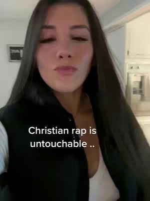 A woman with long black hair, wearing a black bomber jacket over a white top, is the central focus. The background is blurred and indistinct, but appears to be indoors. Text overlayed on the image reads: 'Christian rap is untouchable ..'.