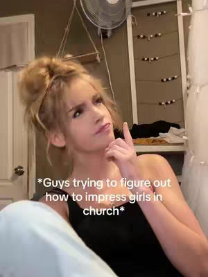 A woman with blonde hair in a bun is shown from the shoulders up. She is wearing a black top and looking upwards with a thoughtful expression. The background is a blurred room with a fan and some shelves.