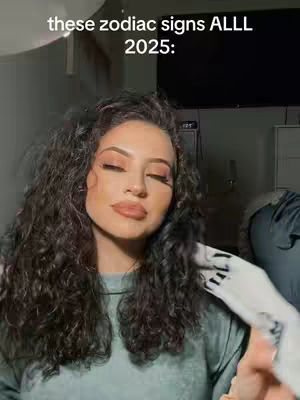 A woman with long, curly dark hair and full makeup, including eyeshadow and lip color, gazes slightly downward with closed eyes. Text overlayed in a stylized white font reads, "these zodiac signs ALLL 2025:" She is wearing a grey top in a dimly lit setting.