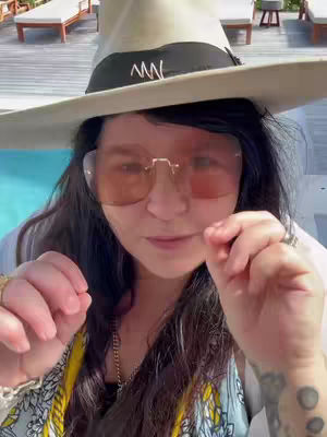 A woman wearing a wide-brimmed hat, sunglasses, and jewelry is the central focus. She is outdoors, with a pool and lounge chairs visible in the background.