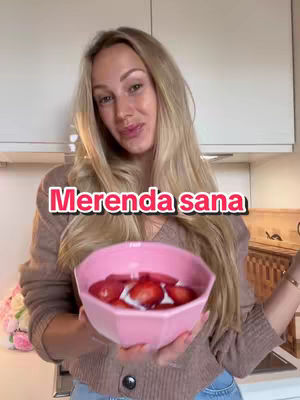 A woman with long blonde hair smiling and holding a pink bowl filled with strawberries sitting in front of a bright kitchen.