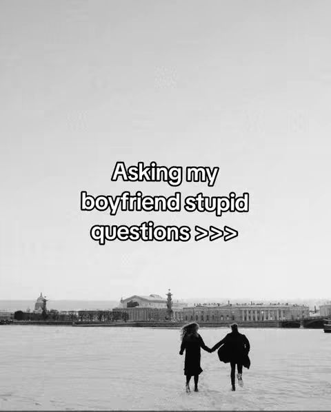 A grayscale photo shows a couple walking hand-in-hand, seen from behind, along a beach or a snow-covered area. In the background, there is a city skyline, with buildings and architecture visible. The sky is bright and clear, and the general mood is peaceful and romantic. The overlay text states, “Asking my boyfriend stupid questions >>>”.