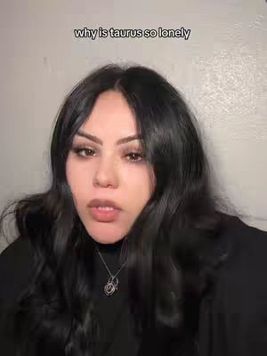 A woman with long, dark hair against a neutral background is the focus. She has a slight smile and is wearing a necklace. Text overlayed at the top reads 'why is taurus so lonely'.