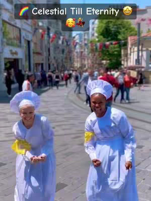 Two women in white church attire are smiling and posing on a city street. The background is a blurred street scene with buildings, people, and flags.