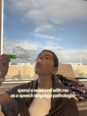 A young woman is blowing bubbles in front of a window with a blue sky and clouds visible outside. The text overlay reads: "spend a weekend with me as a speech language pathologist". Her hair is pulled back, she's wearing a white top and a dark jacket. A bubble wand is in her hand.