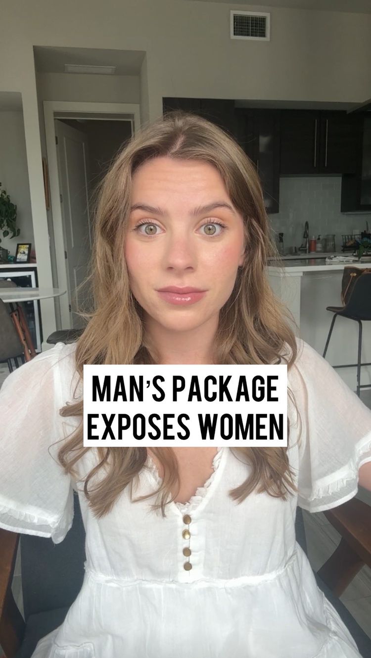 A woman with long, wavy blonde hair is centered, looking directly at the camera with a neutral expression. She wears a white dress with a row of small buttons down the front.  Overlayed at the bottom is a white box with the text 'MAN'S PACKAGE EXPOSES WOMEN'. The background shows elements of a well-lit apartment including a kitchen and neutral-colored walls.