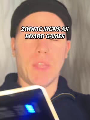 A selfie-style video cover image showing a man wearing a black beanie, looking at the camera with a neutral expression. The background setting is blurred, likely indoors. The text overlay reads 'ZODIAC SIGNS AS BOARD GAMES'. The image has a warm, slightly desaturated color palette.